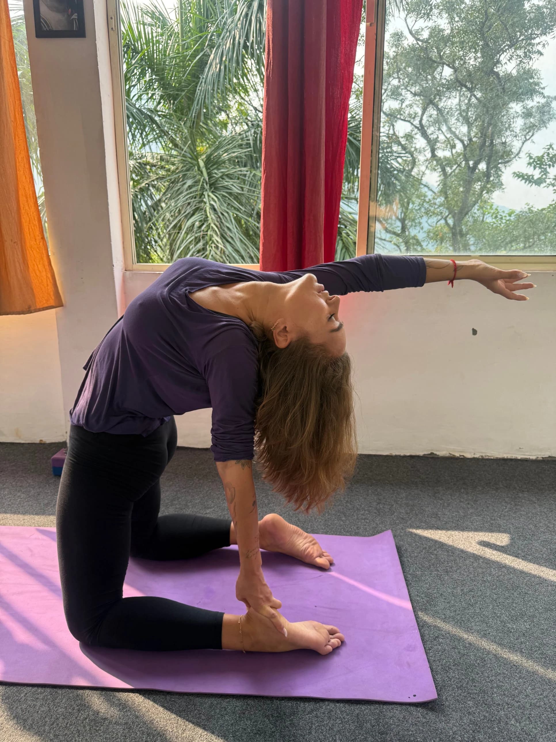 A student in a deep kneeling backbend on a purple mat, by a window with palm trees outside